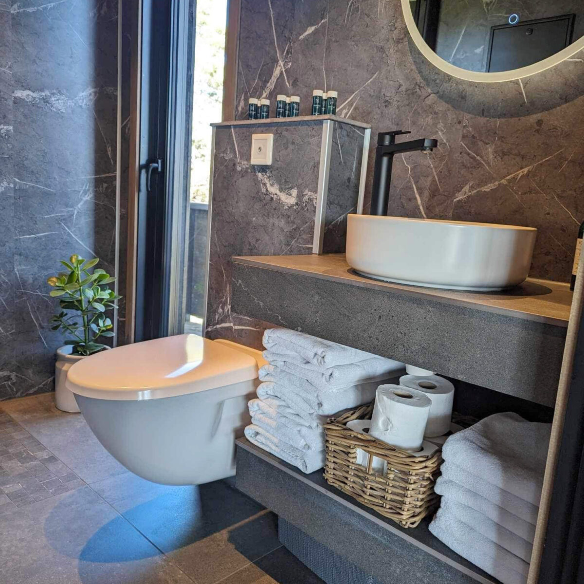 Et moderne baderom med grå marmorerte fliser, stabler med hvite og fint brettede håndduker ligger på benken. Et moderne vegghengt toalett fra Jets.